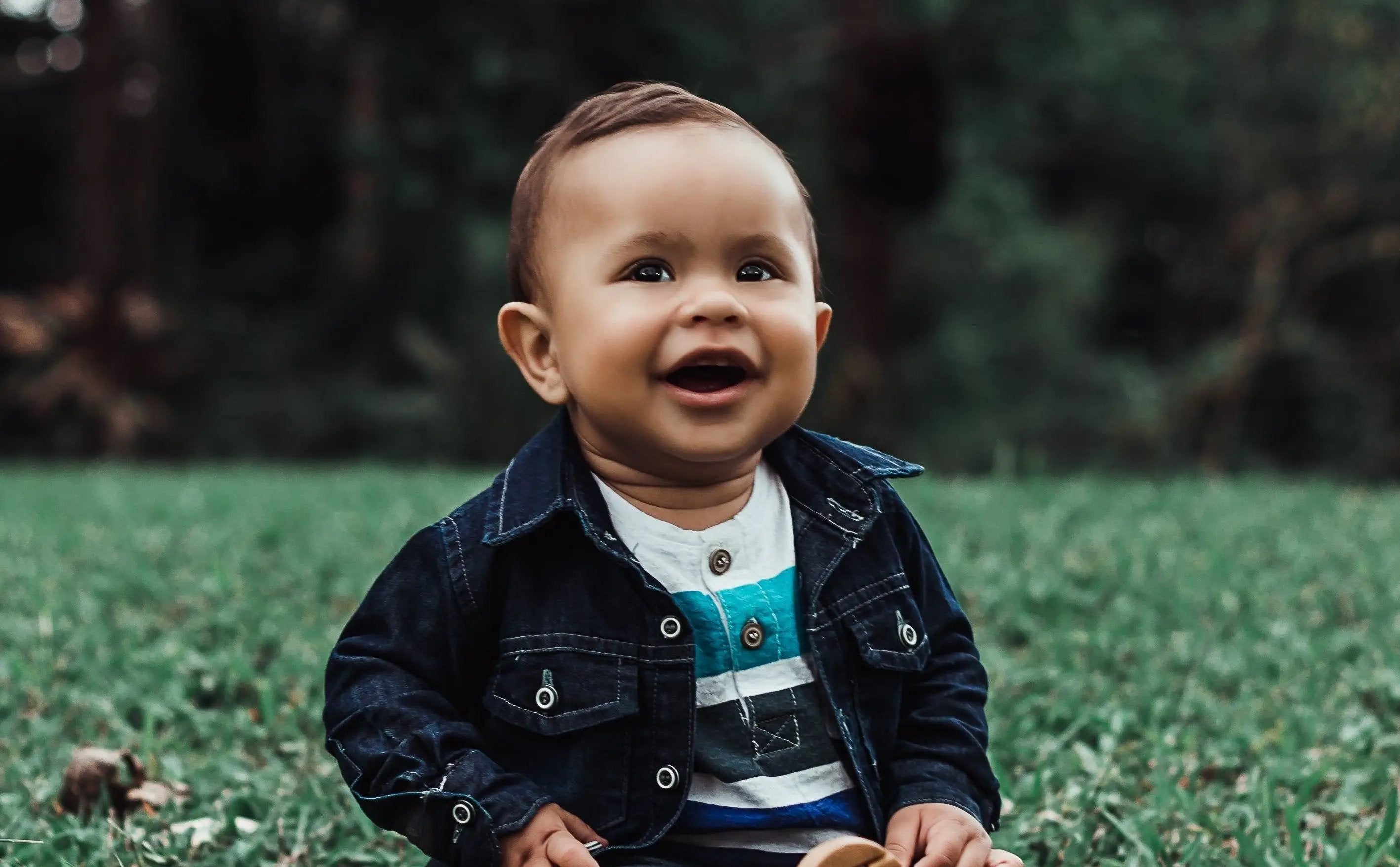 Baby boy smiling on the grass in jacket and stripy shirt.